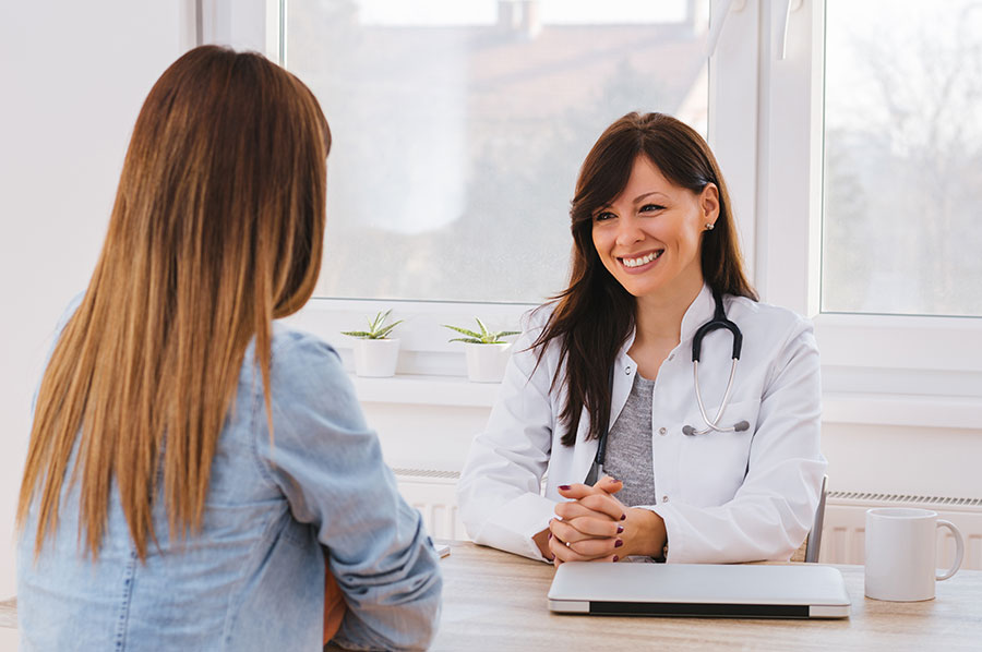 Médica sonriente con bata blanca y estetoscopio atendiendo a una paciente en consulta, imagen representativa de atención personalizada en clínica de cirugía estética Alicante